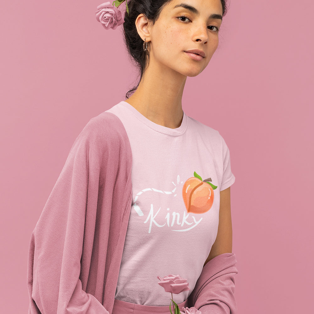 model wearing light pink girly slim fitted t-shirt with short sleeves with a print in a front of a whip hitting a peach and the white text kinky